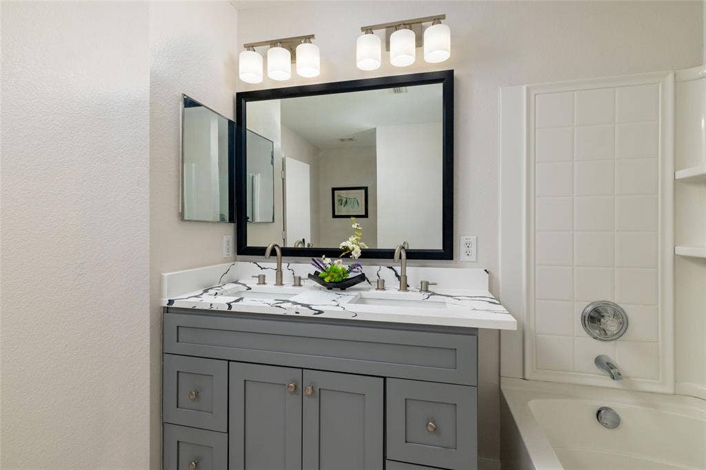 Grey shaker-style dual vanity with a white-and-grey veined countertop, dual undermount sinks, polished nickel faucets, and a framed vanity mirror with dual glass light fixtures