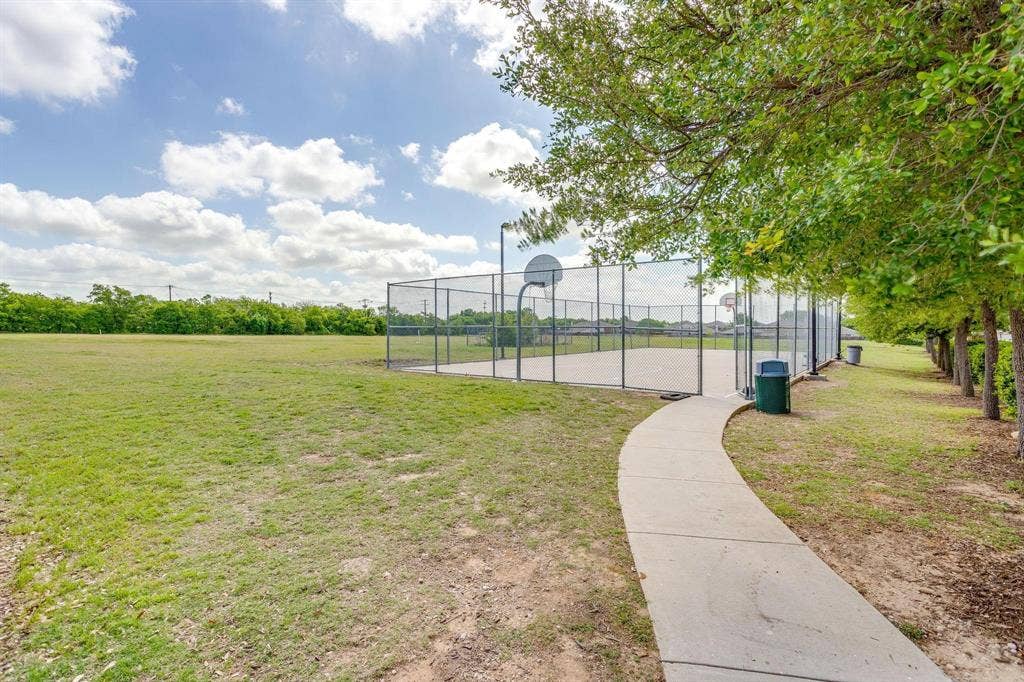 Outdoor recreation area featuring a paved basketball court, chain-link fencing, a concrete walkway, and expansive grassy grounds