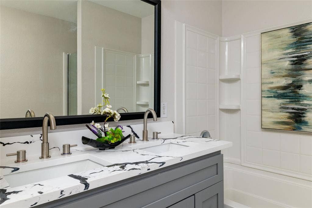 Double-sink vanity featuring a white surface with dark veining, grey shaker-style cabinetry, brushed nickel arched faucets, and a large rectangular mirror with a black frame