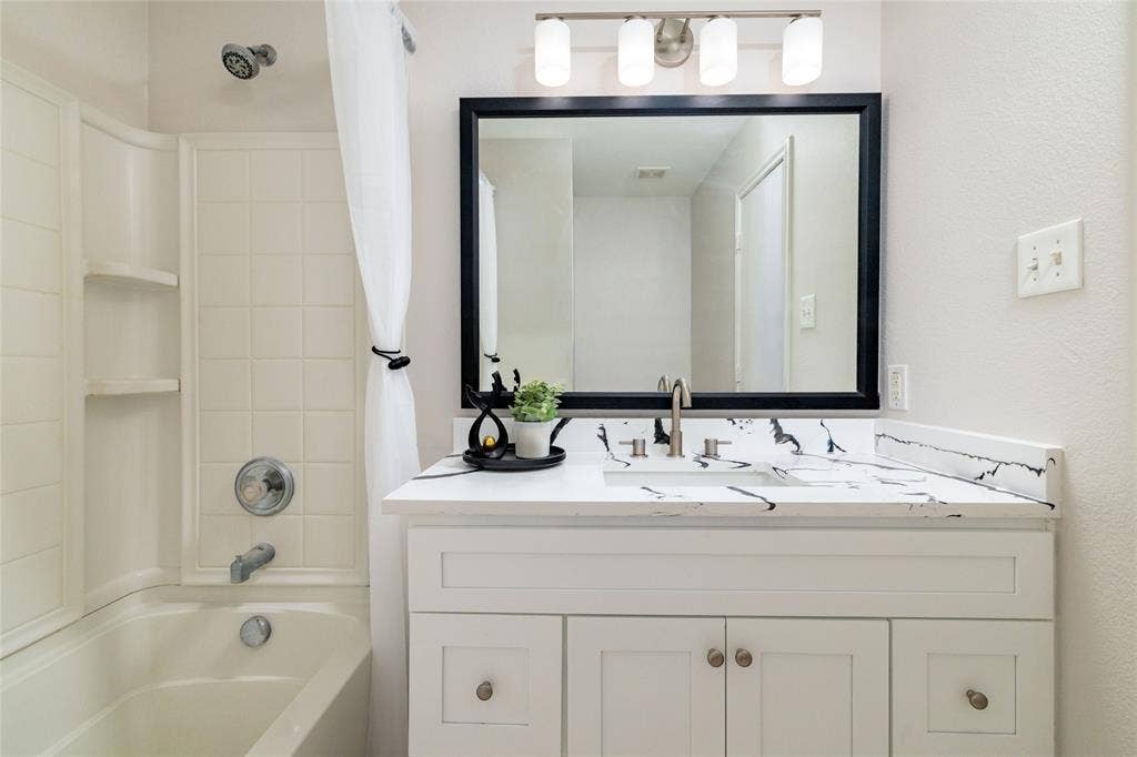 White vanity featuring a contrasting stone-finish countertop with an integrated rectangular sink and polished nickel hardware, complemented by a framed vanity mirror, four-bulb light fixture, and a tile-surround tub with built-in shelving
