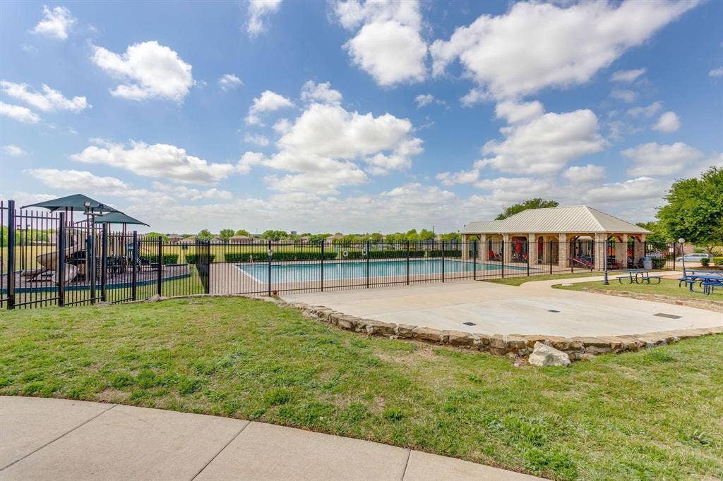 Community swimming pool featuring a concrete deck, metal fencing, adjacent playground equipment, and a covered pavilion with stone-column supports
