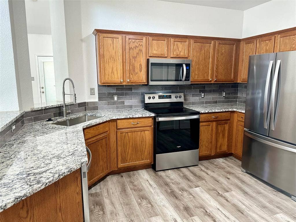 9756 Old Field Drive, McKinney, TX 75072 Kitchen featuring wood cabinetry, stone countertops, gray subway tile backsplash, stainless steel appliances, and wood-finish flooring