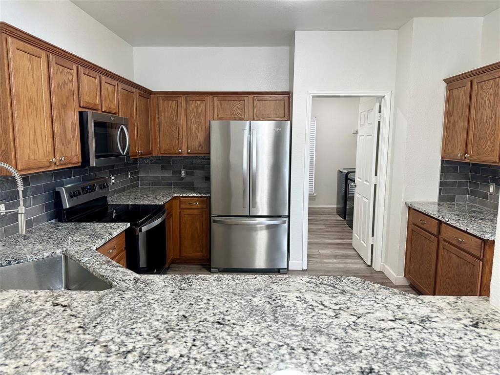 9756 Old Field Drive, McKinney, TX 75072 Kitchen featuring medium-tone wood cabinetry, stone countertops, dark tile backsplash, stainless steel appliances, and a stainless steel undermount sink