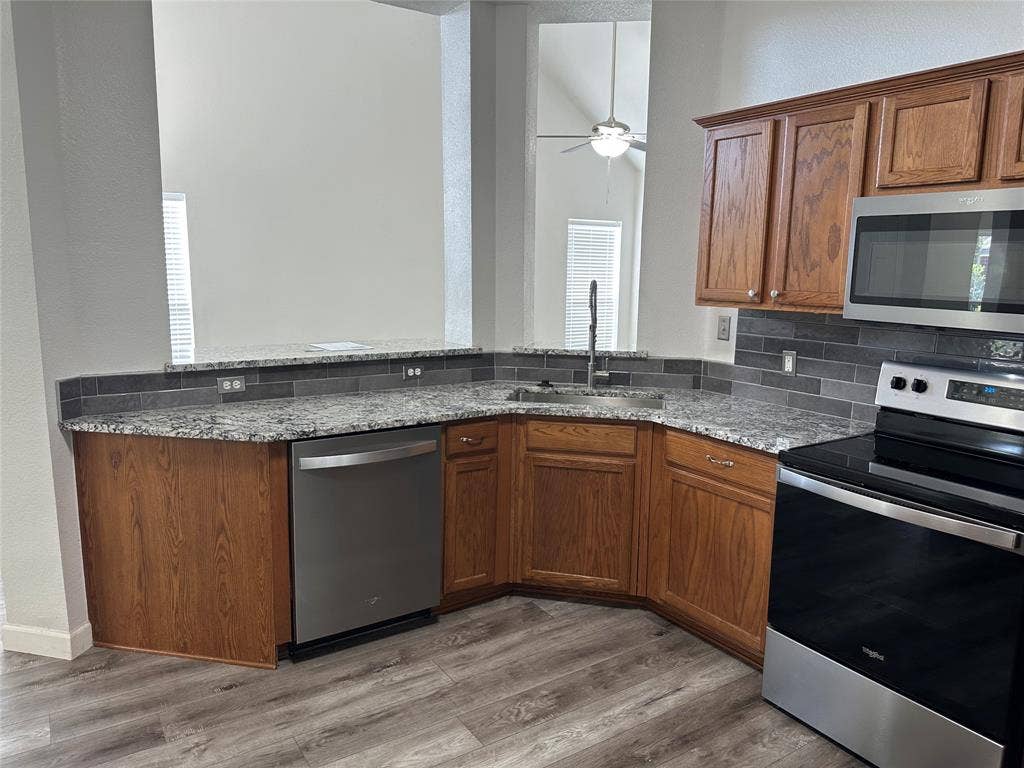 9756 Old Field Drive, McKinney, TX 75072 Kitchen featuring stone-finish countertops, stainless steel appliances, stained wood cabinetry, charcoal tile backsplash, and wood-finish flooring