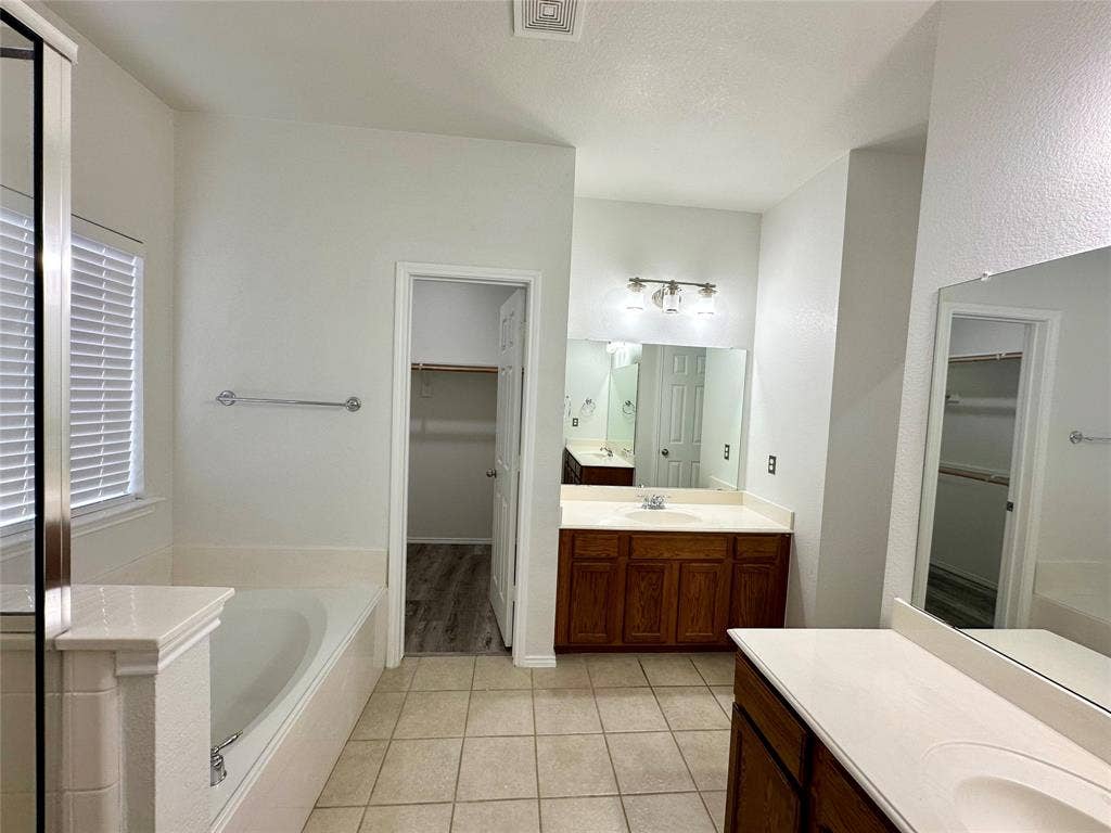 9756 Old Field Drive, McKinney, TX 75072 Bathroom featuring dual vanities with wood-finish cabinetry, solid-surface countertops, integrated sinks, and large wall-mounted mirrors, complemented by a soaking tub with tiled surround and ceramic floor tiling