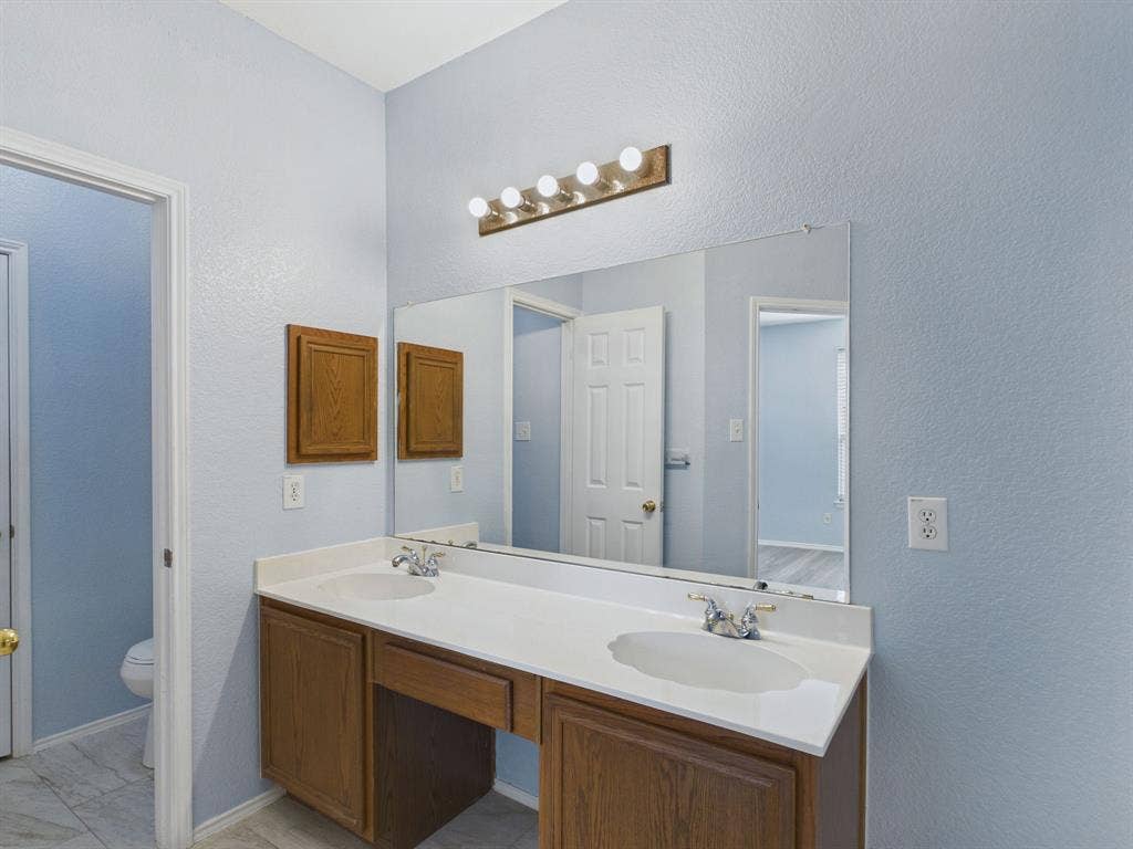 12652 Oakwood Circle, Fort Worth, TX 76040 Double vanity featuring wood-finish cabinetry, a white countertop with integrated dual sinks, chrome fixtures, and a wide-format wall mirror