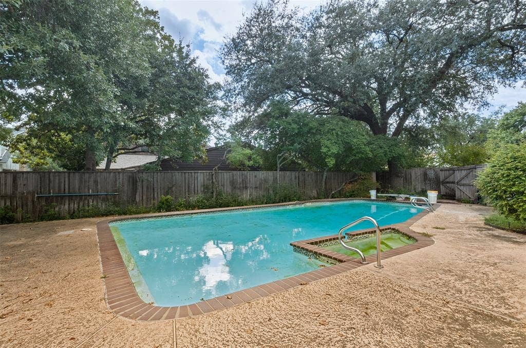 6936 Wildglen Drive, Dallas, TX 75230 In-ground swimming pool featuring a brick-paver border, a dedicated spa area, metal handrails, and a concrete deck enclosed by a wood privacy fence with mature tree canopy