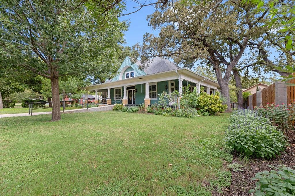 Single-story residence featuring a wrap-around porch with column supports, prominent front gable with decorative trim, exterior lap siding, expansive manicured lawn, and mature canopy trees