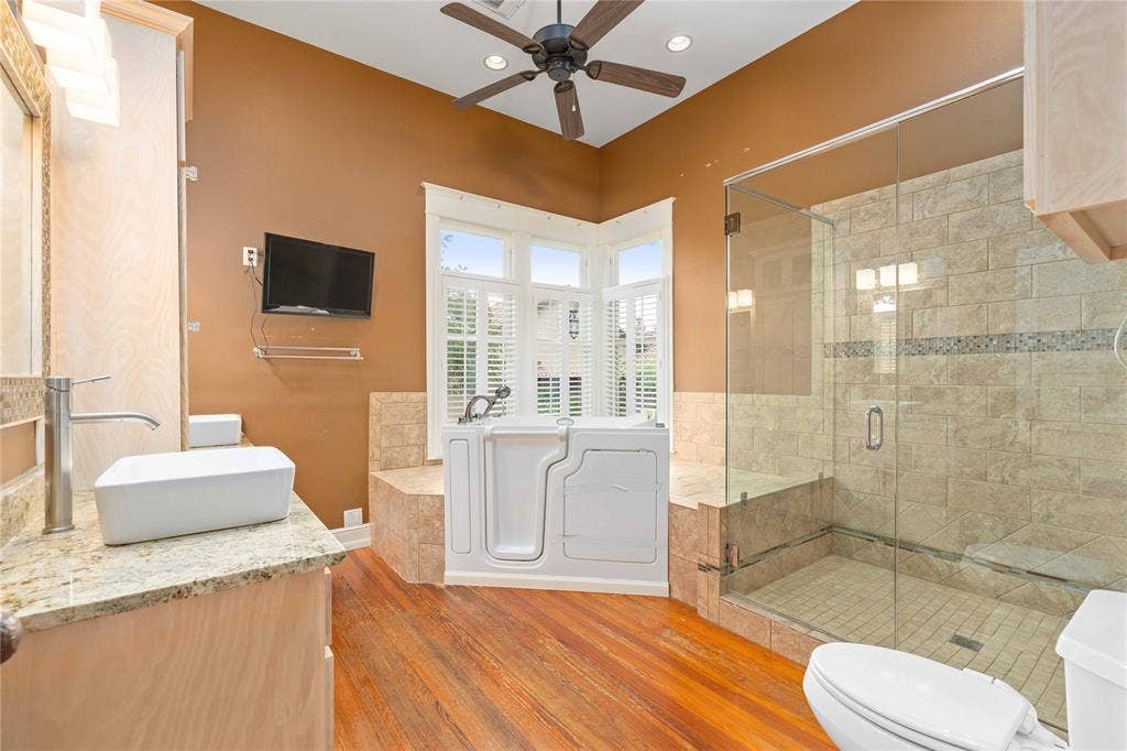 Bathroom featuring wood-finish flooring, a frameless glass-enclosed shower with stone-tile surround, a step-in soaking tub, stone countertop with a rectangular vessel sink, and ceiling-mounted fan