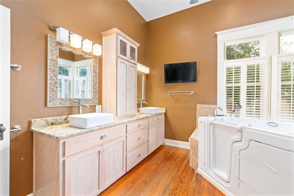 Bathroom featuring light wood-finish cabinetry, a stone-surfaced dual vanity with rectangular vessel sinks, walk-in soaking tub, wood-finish flooring, and plantation shutters