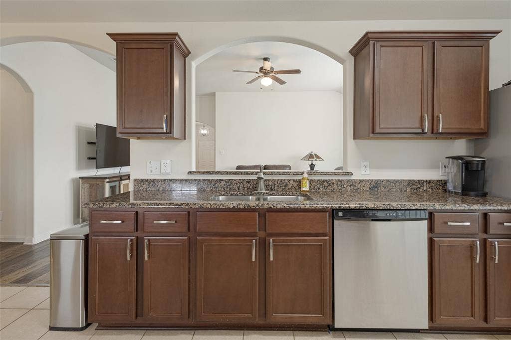 Kitchen featuring dark wood cabinetry, stone countertops, dual-basin stainless steel sink, built-in stainless steel dishwasher, and an arched architectural opening