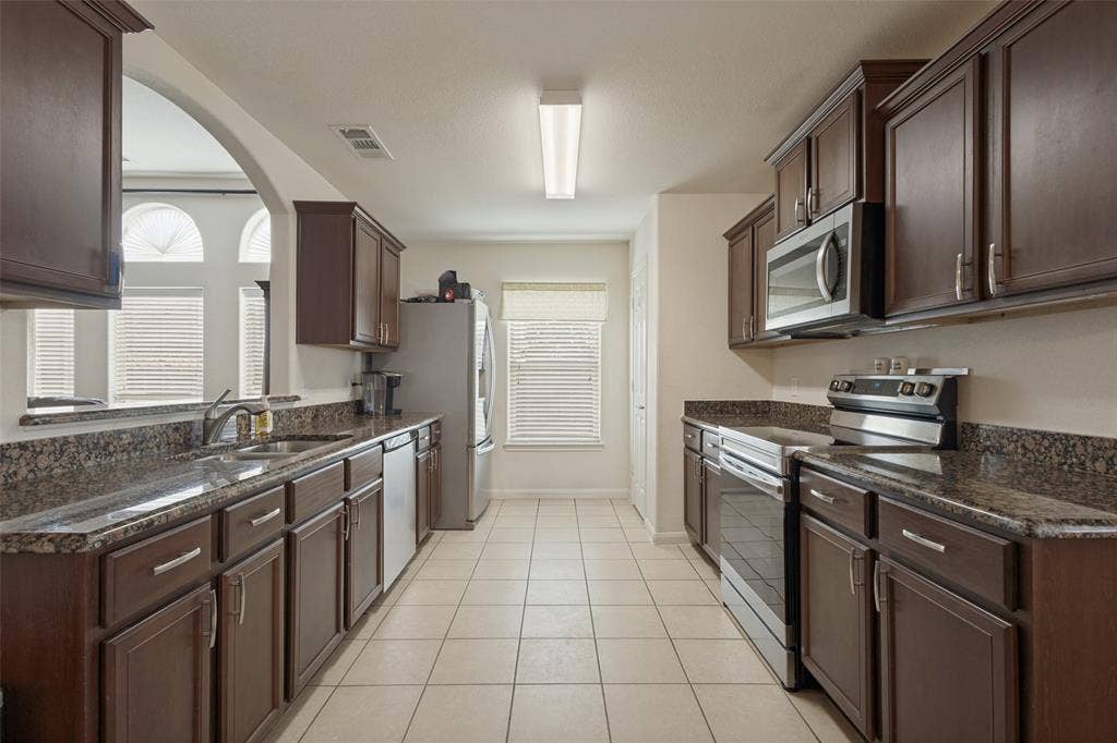 Kitchen featuring dark-finish cabinetry, stone-style countertops, stainless steel appliances, ceramic tile flooring, and a large arched pass-through window