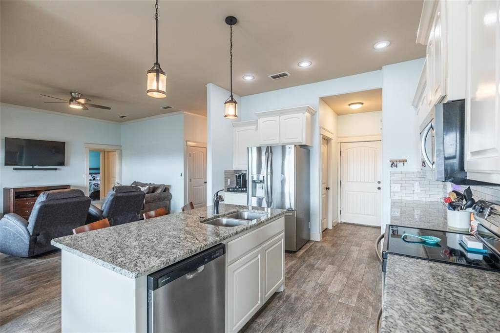 Kitchen featuring a central island with a stone countertop and double-basin sink, white shaker-style cabinetry, stainless steel appliances, pendant lighting, and wood-finish flooring