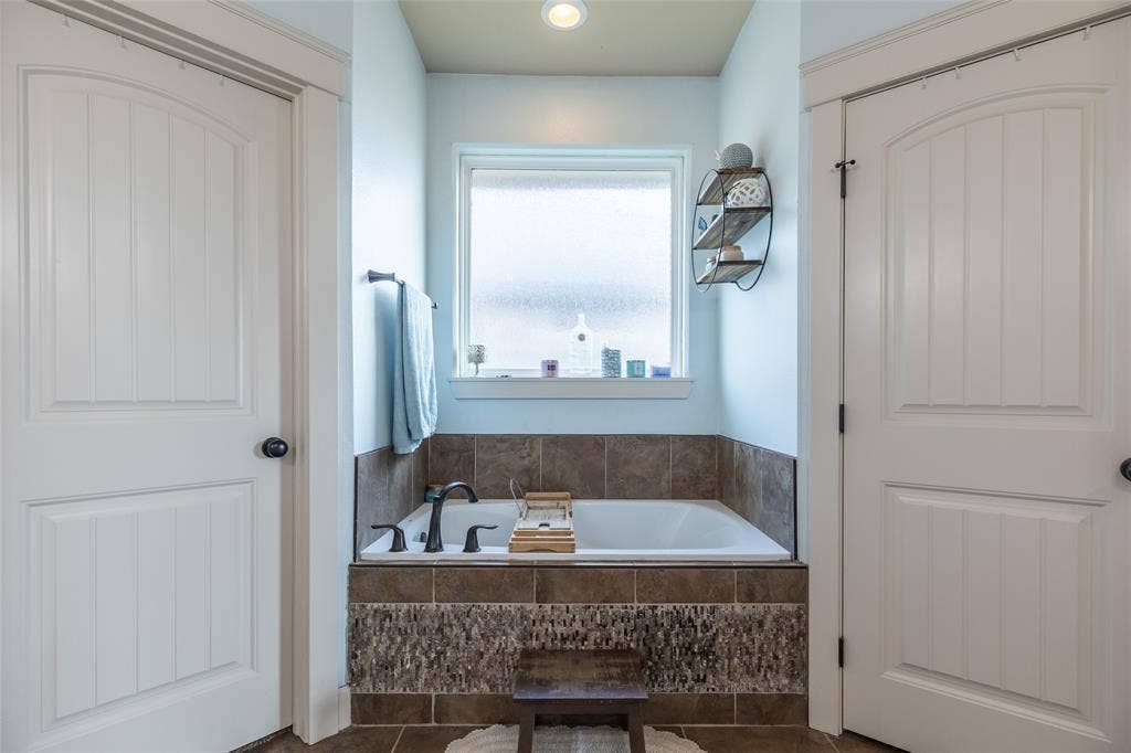 Inset soaking tub featuring a stone-tiled surround, dark metallic faucet fixtures, privacy-glass window, and two-panel interior doors with casing