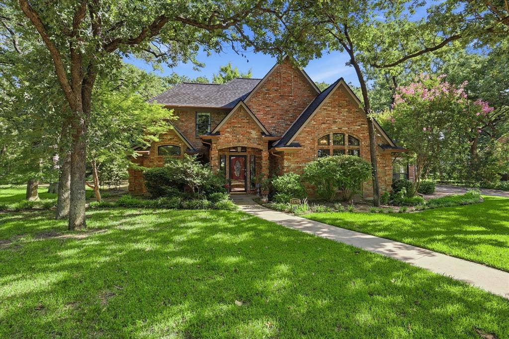 Traditional brick exterior featuring a steeply pitched roof, arched windows, covered entrance, and mature trees on a landscaped lot