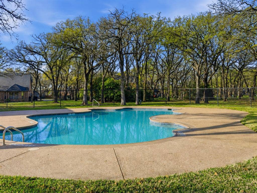 In-ground swimming pool featuring a textured concrete deck, metal perimeter fencing, and mature tree canopy backdrop