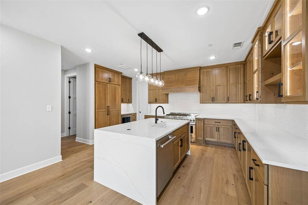 Kitchen featuring custom wood cabinetry, an expansive center island with quartz countertops, integrated professional-grade gas range, wood-finish flooring, and linear multi-pendant lighting