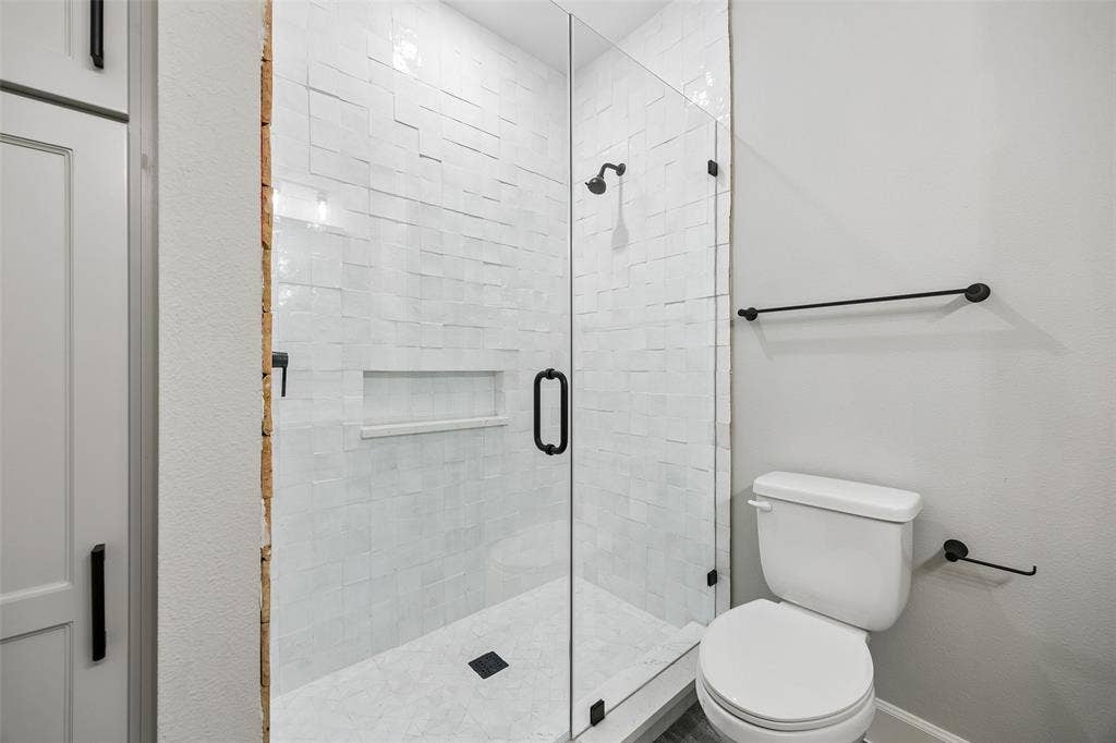 Walk-in shower featuring textured white tile walls, built-in recessed niche, frameless glass door, and matte black fixtures, paired with a coordinating toilet and matte black hardware