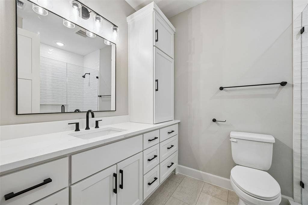 White cabinetry with matte black hardware, stone-surface countertop, rectangular undermount sink, built-in storage tower, and a framed mirror with integrated lighting