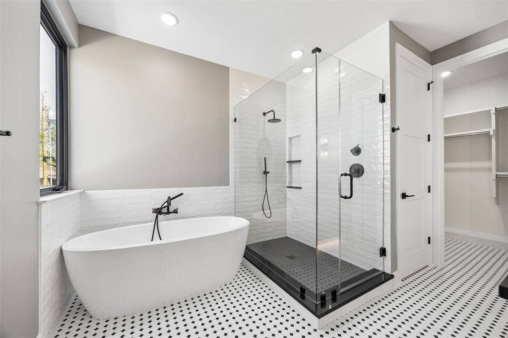 Freestanding soaking tub with floor-mounted faucet, glass-enclosed shower with dark tiled base and recessed shelving, and patterned mosaic tile flooring