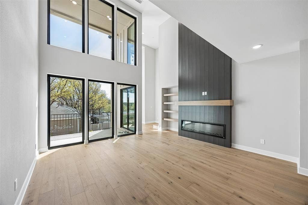 Double-height living area featuring floor-to-ceiling windows, wide-plank wood-finish flooring, a vertical board-accented fireplace surround, a built-in horizontal mantel, and recessed shelving