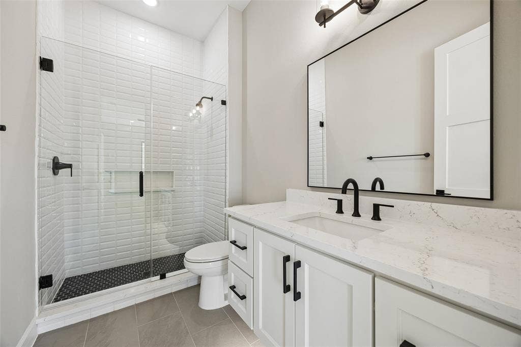 Bathroom featuring a walk-in shower with white subway tile walls and dark patterned flooring, light-toned cabinetry with a solid surface countertop, an undermount sink, and matte black fixtures