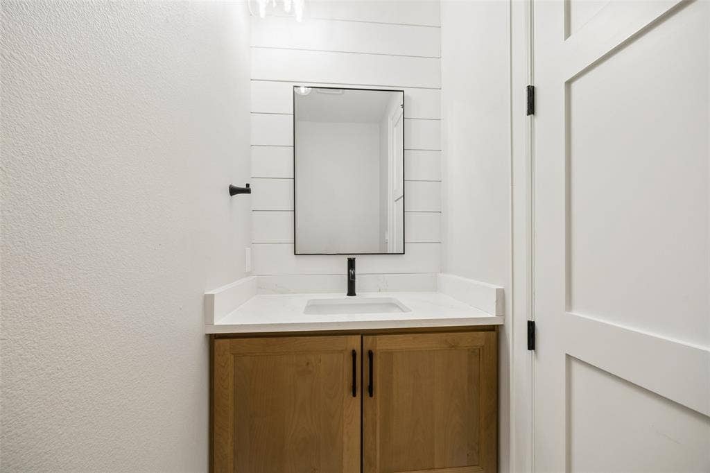 Vanity featuring a light wood-finish cabinet, white countertop with an integrated rectangular sink, shiplap accent wall, framed mirror, and matte black hardware