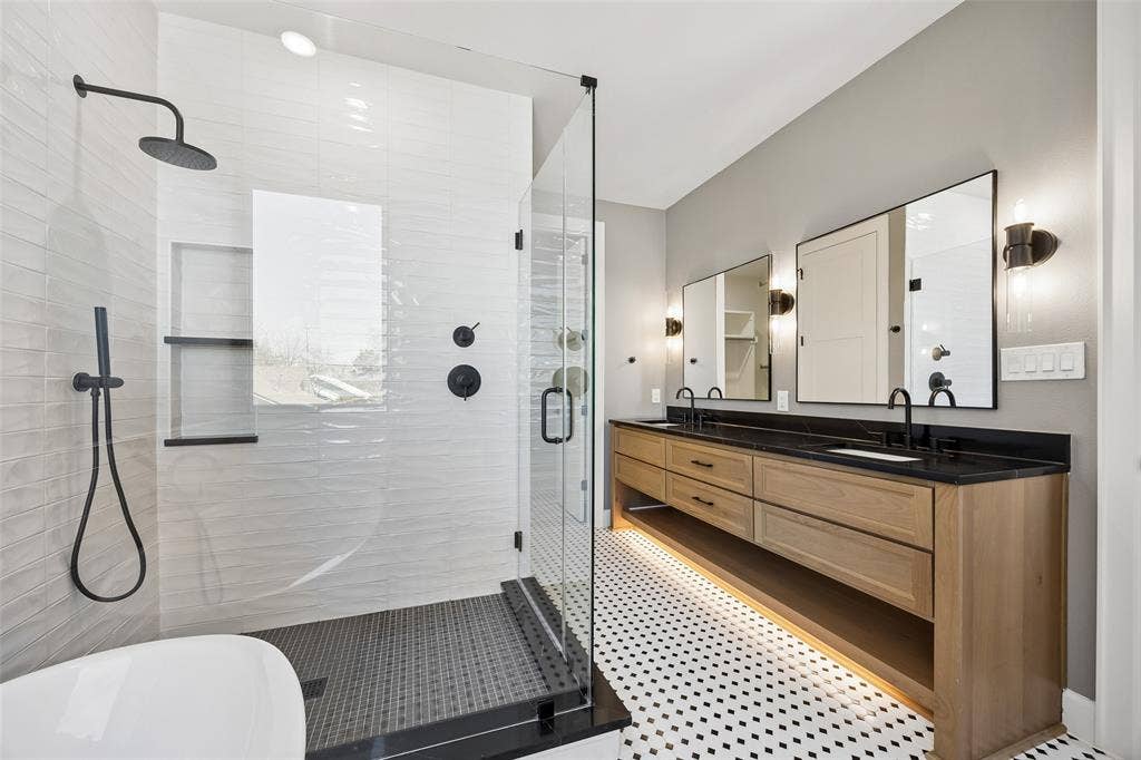 Walk-in shower with textured tile surround, built-in shelving, glass enclosure, and matte black hardware; double vanity with wood-finish cabinetry, dark solid-surface countertop, and under-cabinet lighting