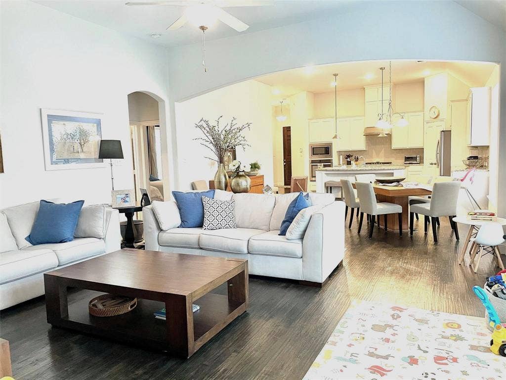 Open-concept living space featuring wood-finish flooring, vaulted ceilings, and a large arched entryway leading to a kitchen with white cabinetry and a central island