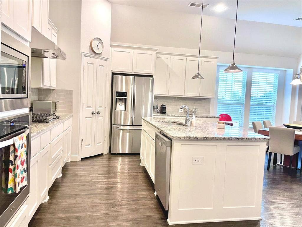 Kitchen featuring white cabinetry, stone countertops, stainless steel appliances, a central island with an integrated sink, and wood-finish flooring