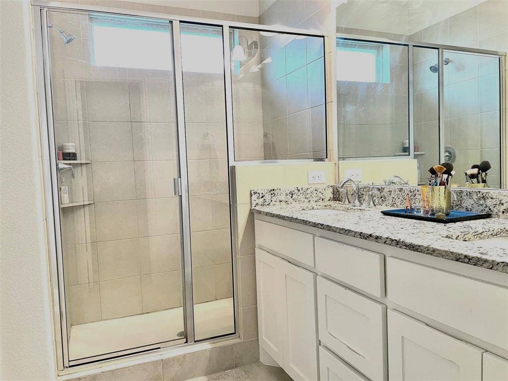 Shaker-style white cabinetry, speckled stone countertop, dual-basin vanity, and a tiled shower enclosure with a glass door