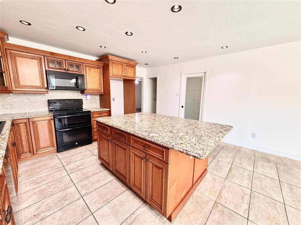 Kitchen featuring wood cabinetry, stone-finish countertops, a central island, black appliances, and tile flooring