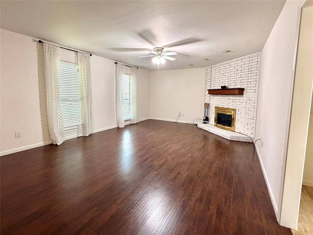 Spacious living area featuring wood-finish flooring, a white-painted brick fireplace, a wooden mantel, and a central ceiling fan