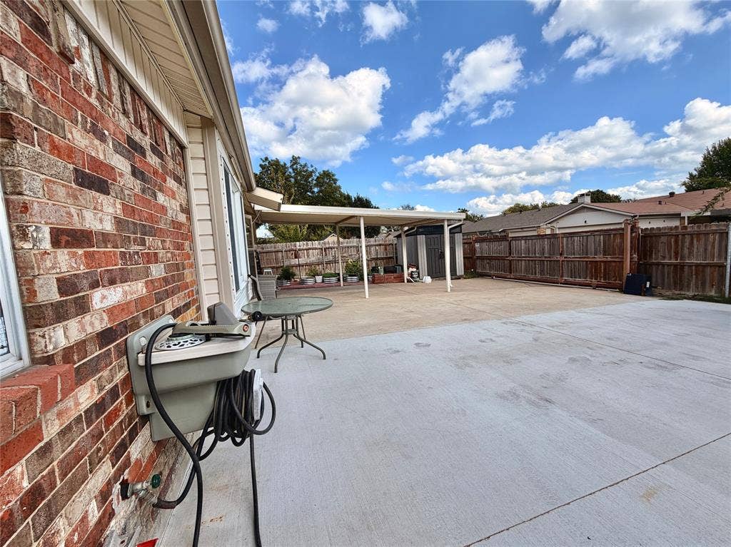 Expansive concrete patio area featuring a covered carport, full-perimeter wood privacy fencing, and exterior brick masonry