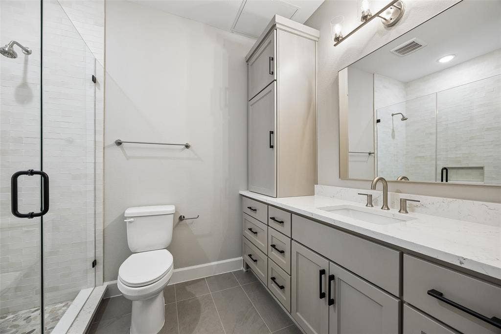 Bathroom featuring a vanity with neutral-toned cabinetry, stone-look countertop, integrated sink, and matte black hardware, accompanied by a glass-enclosed shower with tiled surround, full-width mirror, and built-in vertical storage cabinet