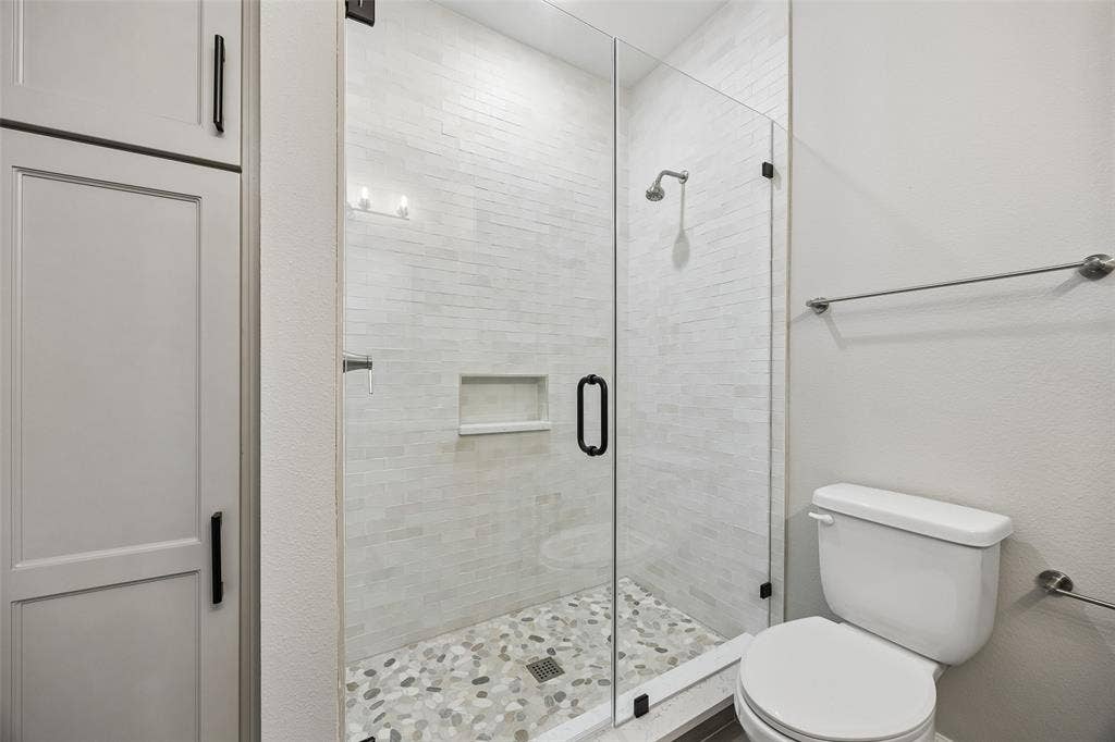 Walk-in shower featuring floor-to-ceiling tile, integrated recessed shelving, a frameless glass door, and a pebble-stone basin floor, complemented by floor-to-ceiling cabinetry with matte black hardware