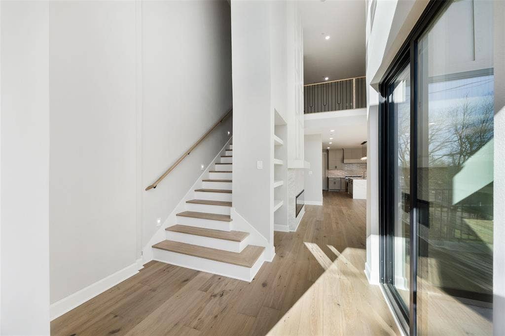 Double-height ceiling, wood-finish flooring, staircase with a wood-finish handrail, built-in wall shelving, and floor-to-ceiling glass sliding doors