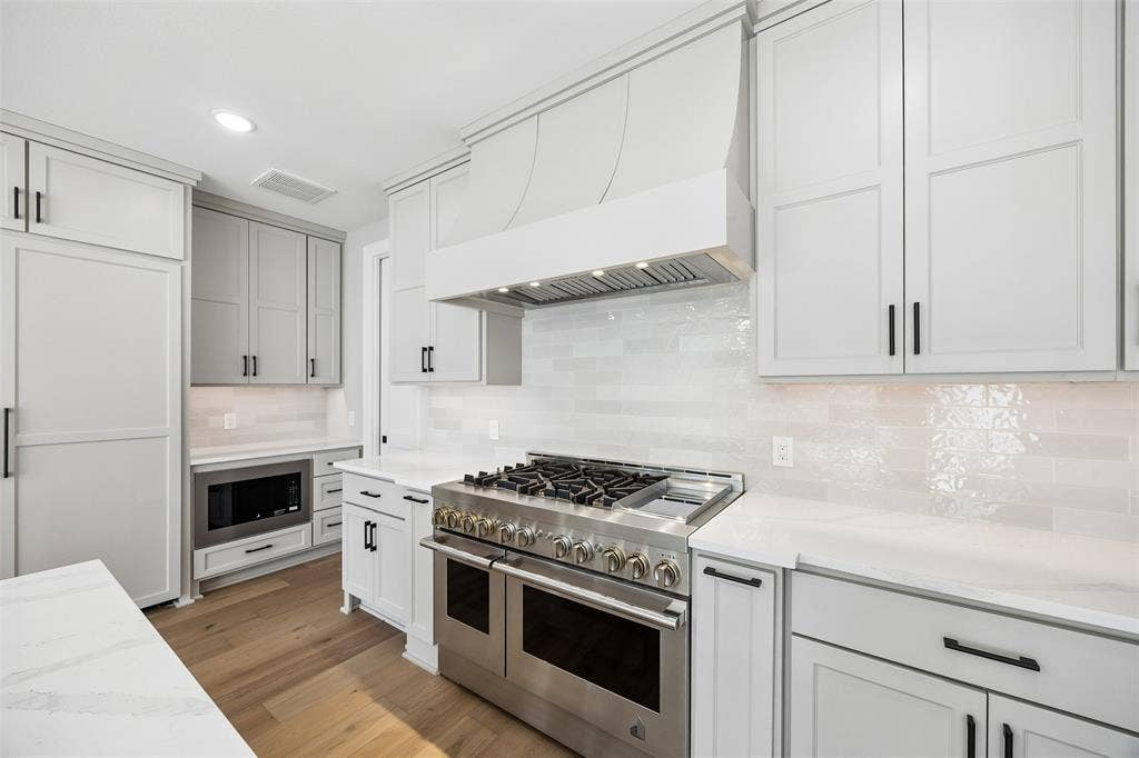 Kitchen featuring custom shaker-style cabinetry, professional-grade stainless steel range with integrated hood, light-toned stone-finish countertops, textured tile backsplash, and wood-finish flooring
