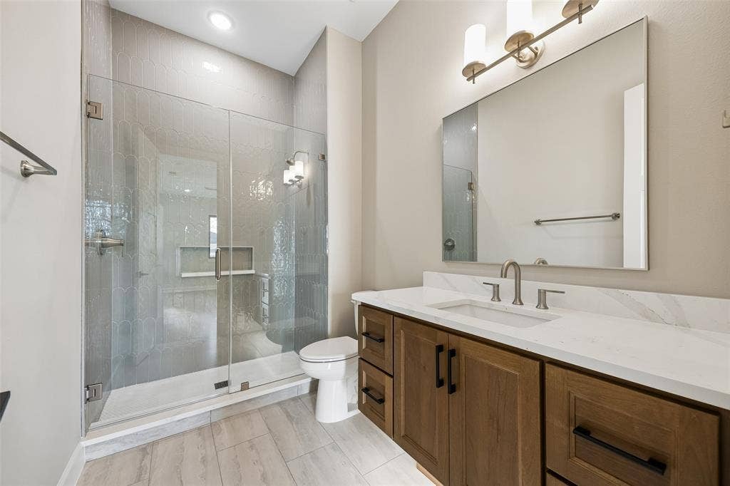 Bathroom featuring a wood-finish vanity with a stone-look countertop, an integrated undermount sink, a frameless glass-enclosed shower with a recessed niche, and wood-finish flooring