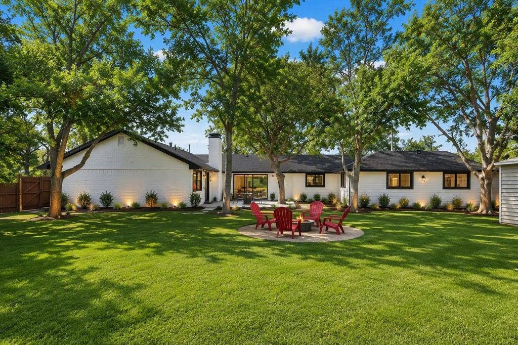 Expansive backyard featuring a circular stone fire pit area, manicured lawn, mature trees, and a painted brick exterior with black trim