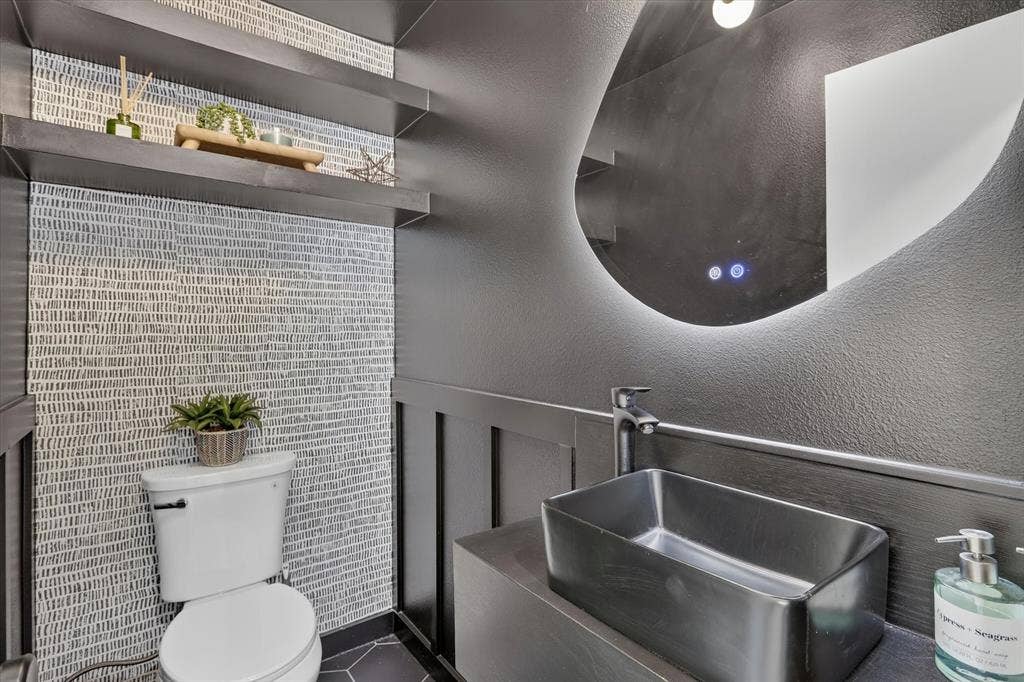 Powder room featuring patterned wall covering, board and batten wainscoting, matte black vessel sink with high-arc faucet, floating shelves, and illuminated frameless mirror