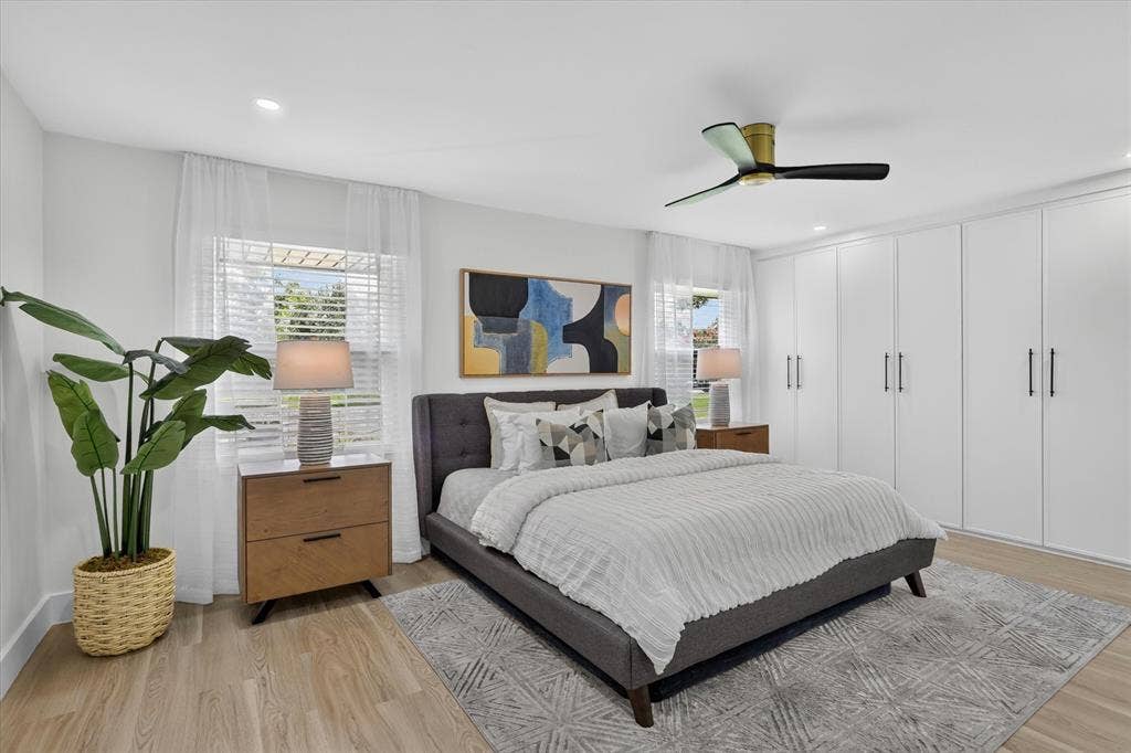 Full-height wall-to-wall white cabinetry with matte black hardware, wood-finish flooring, dual windows with horizontal blinds, and a modern ceiling fan with matte black blades
