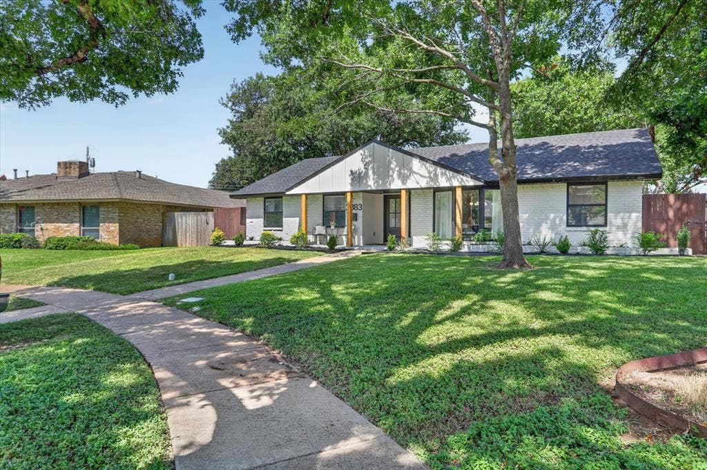 Single-story exterior featuring a painted brick facade, gabled roofline, wood-finish porch columns, and expansive front lawn with mature trees