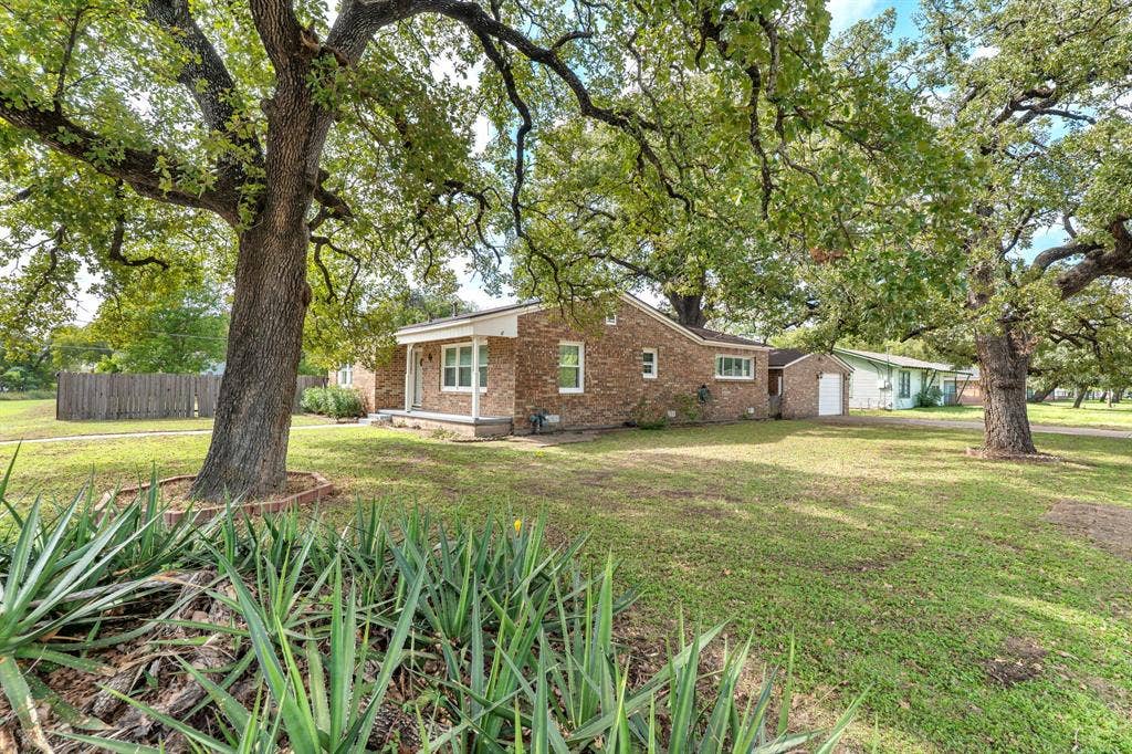 920 Avenue C, Lacy Lakeview, TX 76705 Single-story brick exterior featuring a covered front porch, an attached garage, and mature trees on a lot with perimeter wood fencing