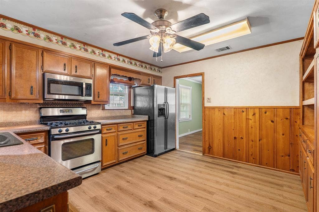 920 Avenue C, Lacy Lakeview, TX 76705 Kitchen featuring wood-finish cabinetry, stainless steel appliances, wood-finish flooring, half-wall wood paneling, and a ceiling fan with integrated lighting