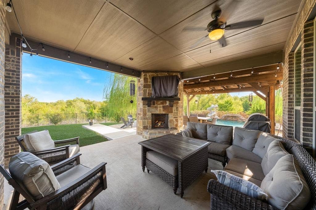 Covered patio featuring a stone corner fireplace, tongue-and-groove ceiling, and ceiling fan, with brick exterior pillars and direct access to an outdoor pool and manicured lawn