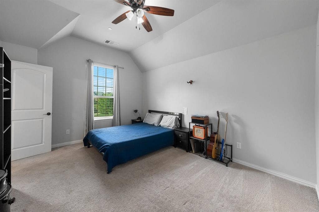 Vaulted ceiling featuring a central multi-blade ceiling fan with integrated light fixture, neutral wall-to-wall carpeting, and a single-pane window with grid detailing