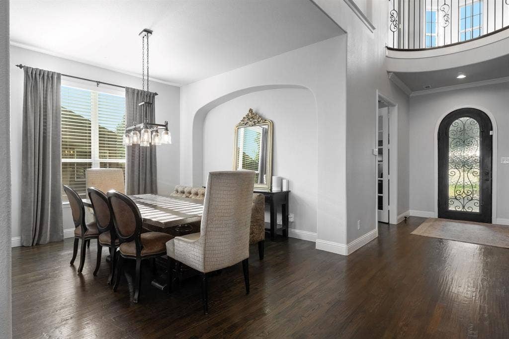 Dining area featuring arched wall detailing, wood-finish flooring, and a multi-light pendant fixture, complemented by an entry featuring an arched door with iron decorative insets and a curved balcony overlook