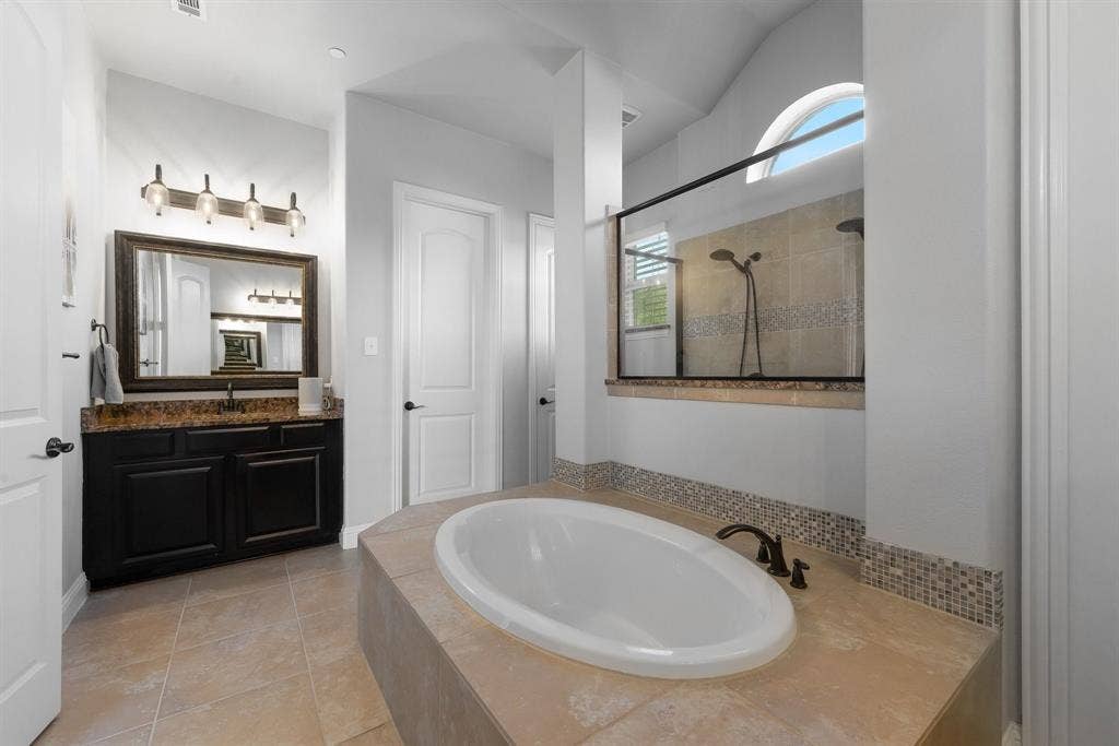 Master built-in soaking tub with tile surround, dark wood vanity, stone countertop, arched window, and glass-enclosed shower