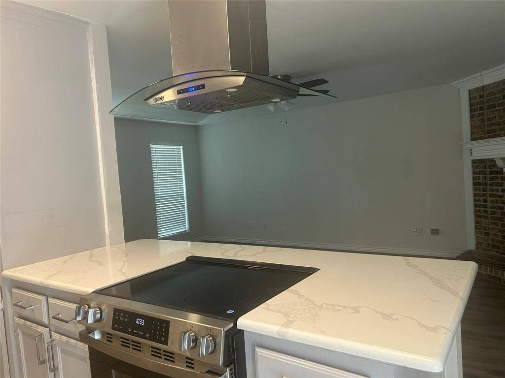 1314 Frio Lane, Garland, TX 75040 Kitchen island featuring a stone-surface countertop, stainless steel electric range, and overhead glass-canopy range hood, complemented by a brick-accent fireplace surround